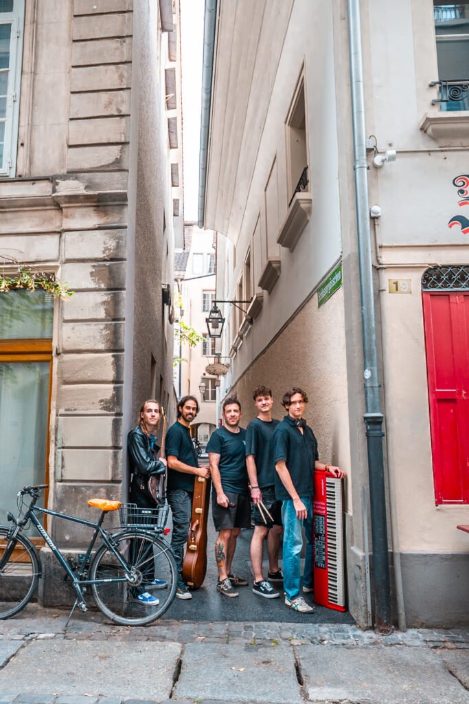 Die fünf Mitglieder von Purple Iris stehen quer, hintereinander gereiht in einer Seitengasse. Die erste Person links trägt eine Bassgitarre. Die zweite Person von Links hält einen Gitarrenkoffer vor sich. Die mittlere Person hält ein Mikrofon in der rechten Hand. Die zweit letzte Person trägt zwei Schlagzeugschläger. Die Person ganz recht hält ein rotes Keyboard das vertikal auf den Boden gestellt ist. 
Links im Bild ist ein Velo zu sehen